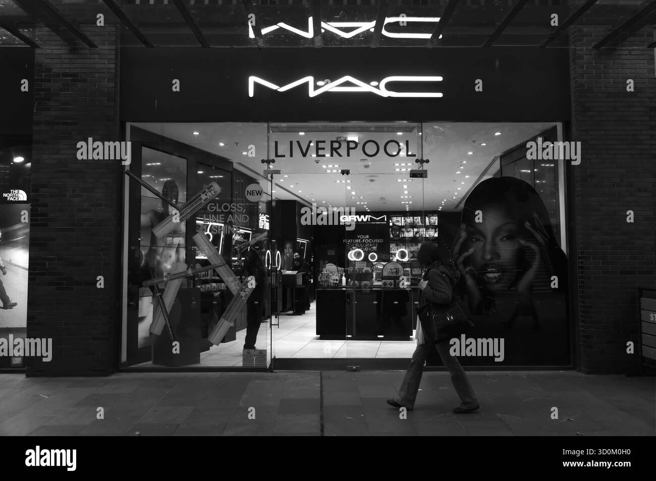 Schwarz-weiß-Foto Einer Frau, die am beleuchteten MAC Cosmetics Store in Liverpool One vorbeigeht, mit hellen Schildern und Make-up-Displays im Inneren. Stockfoto