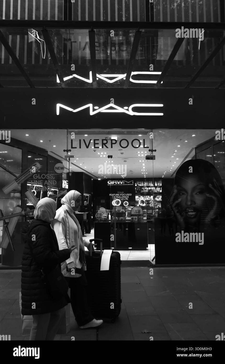 Schwarzweiß-Foto von zwei Frauen, die am beleuchteten MAC Cosmetics Store in Liverpool One vorbeilaufen, mit hellen Schildern und Make-up-Displays im Inneren. Stockfoto
