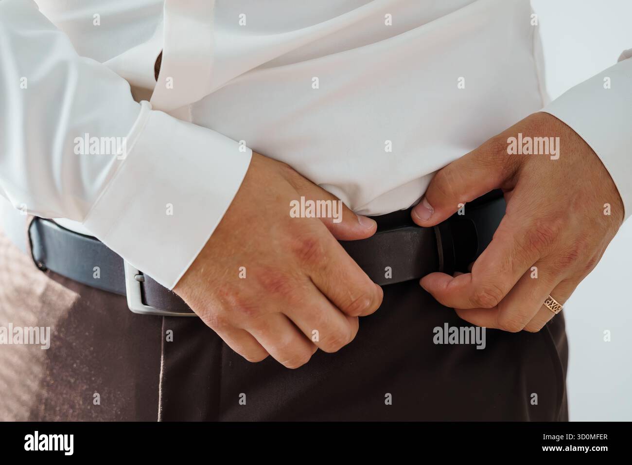 Ein Nahbild zeigt einen Mann, der einen dunkelbraunen Ledergürtel mit einer silbernen Schnalle um die Taille über einer grauen Hose hält, was auf ein formelles OR hindeutet Stockfoto