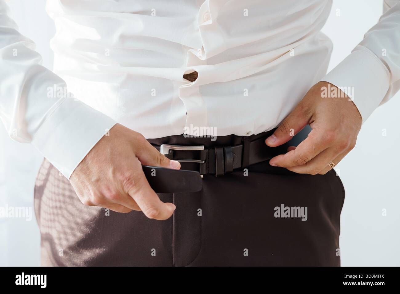 Eine Nahaufnahme zeigt einen Mann, der einen dunklen Ledergürtel um seine Taille hält, braune Hosen und ein helles Hemd trägt, was auf formelle oder bu hindeutet Stockfoto