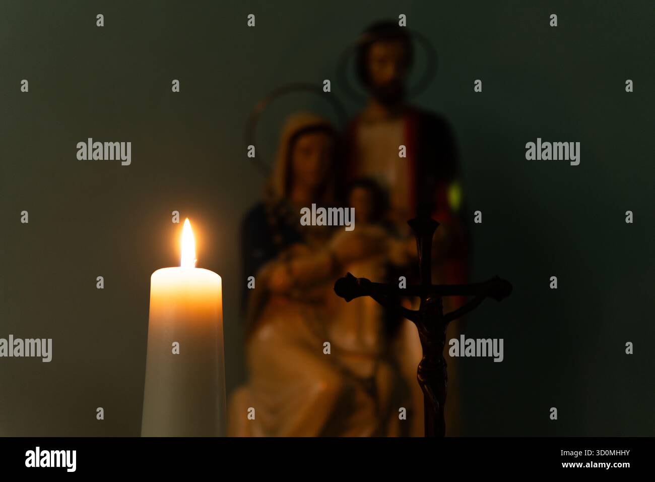 Ein feierliches Foto einer zündeten Kerze, die ein hölzernes Kruzifix beleuchtet. Im weichen, verschwommenen Hintergrund befindet sich eine Statue der Heiligen Familie (Jesus, Maria, Joseph). Stockfoto