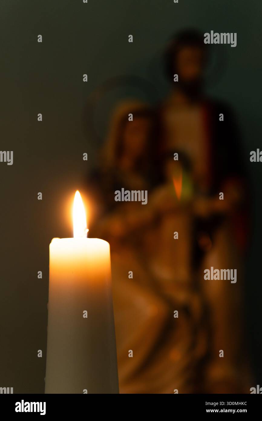 Ein feierliches Foto einer zündeten Kerze, die ein hölzernes Kruzifix beleuchtet. Im weichen, verschwommenen Hintergrund befindet sich eine Statue der Heiligen Familie (Jesus, Maria, Joseph). Stockfoto