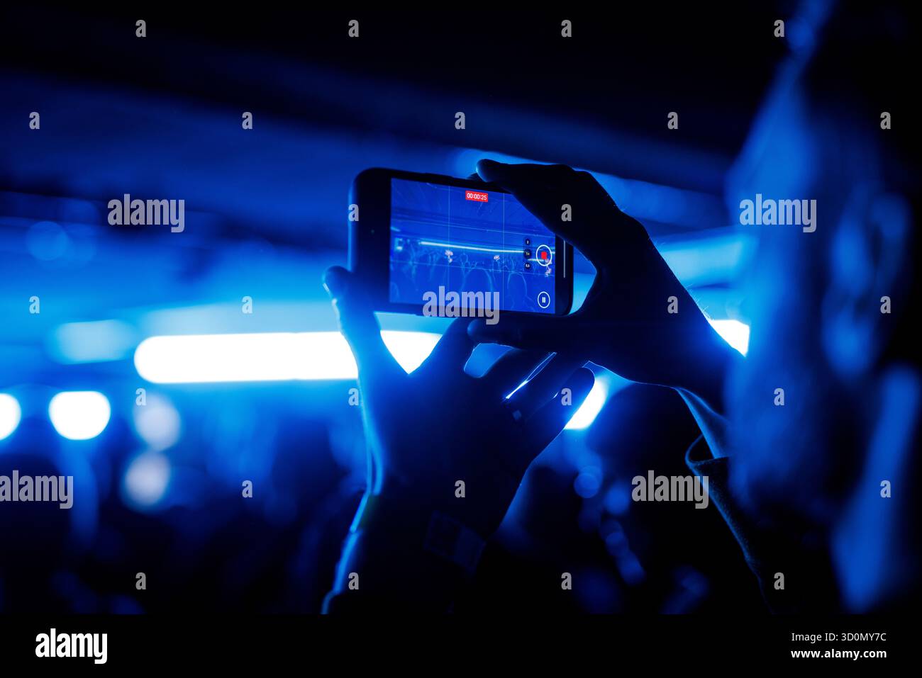 Nahaufnahme der Hände, die das Smartphone halten, während sie das Konzert unter blauem Bühnenlicht filmen. Stockfoto