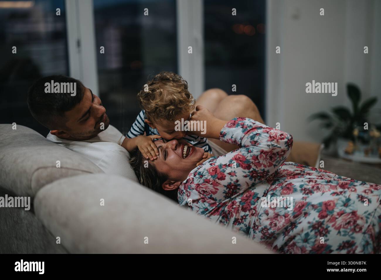 Die Familie genießt einen verspielten Moment auf dem Sofa, während das Kind die Mutter mit Lachen kitzelt Stockfoto