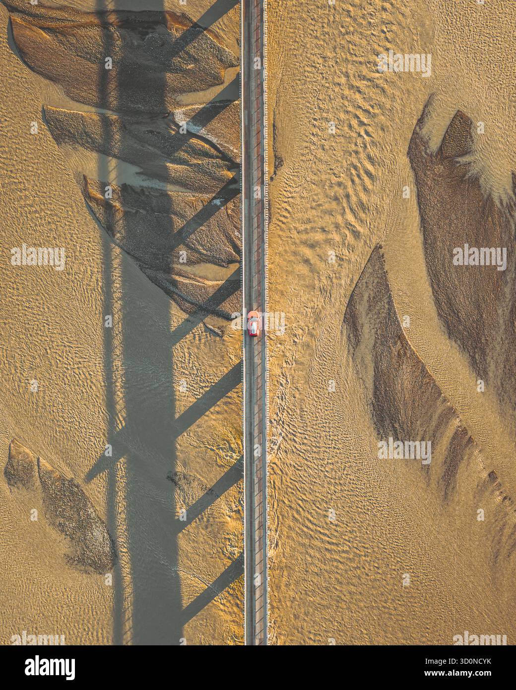 Aus der Vogelperspektive eines einsamen roten Autos, das eine lange Brücke über strukturiertem, sandigem Gelände unter starken Schatten überquert, Stafafafell, Sveitarfélagið Hornafjörður, Island. Stockfoto