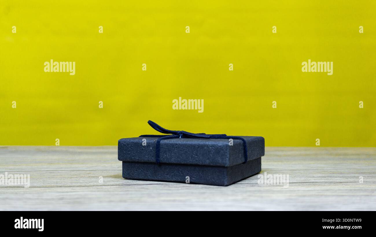 Geschlossene schwarze Geschenkbox auf Holztisch mit hellgelbem Hintergrund, perfekt für festliche, luxuriöse oder Produktpräsentationskonzepte. Stockfoto