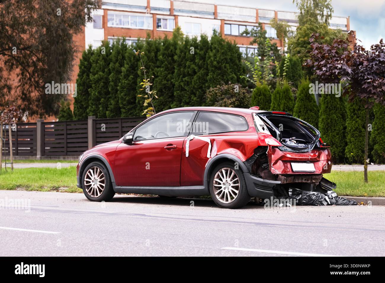 Ein beschädigtes rotes Auto parkt am Straßenrand. Die Rückseite ist zerquetscht, und die Szene zeigt grüne Bäume und ein Wohngebäude im Hintergrund. Stockfoto