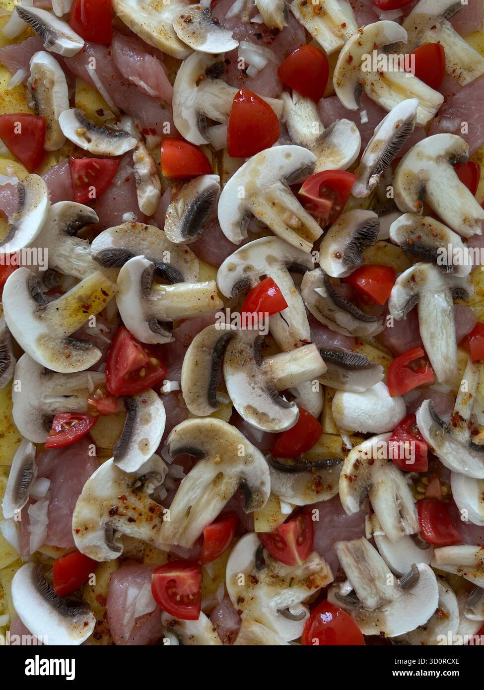 Das Bild zeigt eine Nahaufnahme von rohen geschnittenen Pilzen, gehackten Tomaten, Kartoffeln und Fleischstücken, die gewürzt und für den Ofen zubereitet wurden. Das Foto i Stockfoto
