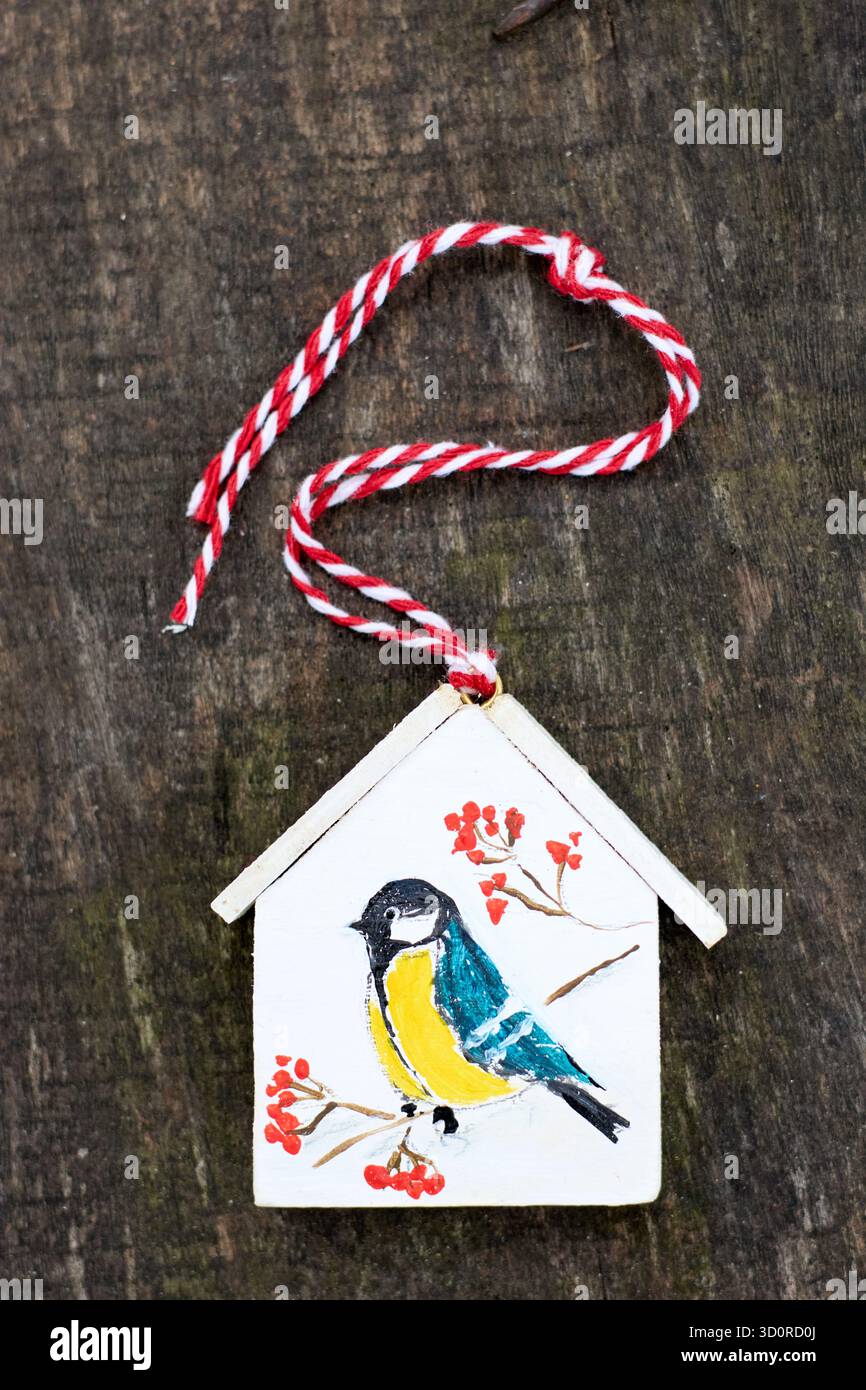 Ein bezauberndes, handgefertigtes Vogelhaus-Ornament mit einem farbenfrohen Vogelgemälde, das von einer rot-weißen, verdrehten Schnur auf einer rustikalen Holzoberfläche aufgehängt wird. Stockfoto