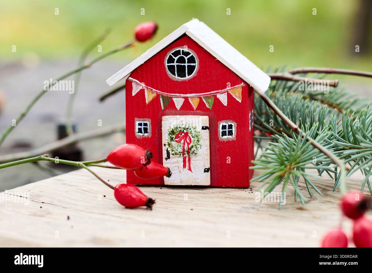 Nahaufnahme eines bezaubernden roten Miniaturhauses mit festlichen Dekorationen, ergänzt durch leuchtende Hüftrosen und üppige Fichtenzweige. Stockfoto