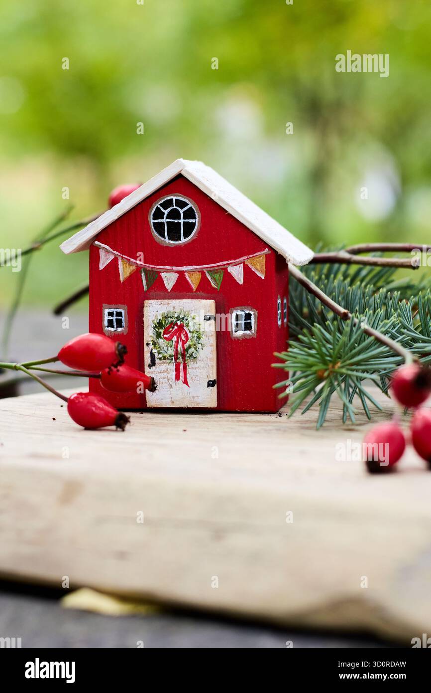 Ein festliches rotes Miniaturhaus mit weißem Dach und einem Kranz an der Tür inmitten roter Beeren und grüner Pflanzen, das einen gemütlichen Urlaubsgeist einfängt. Stockfoto
