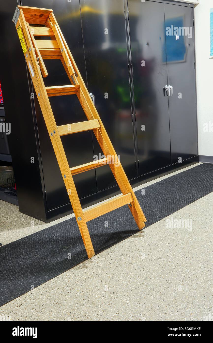 Eine zusammengefaltete Holzleiter oder Trittleiter neigt sich gegen einen schwarzen Schrank in der Garage. Stockfoto