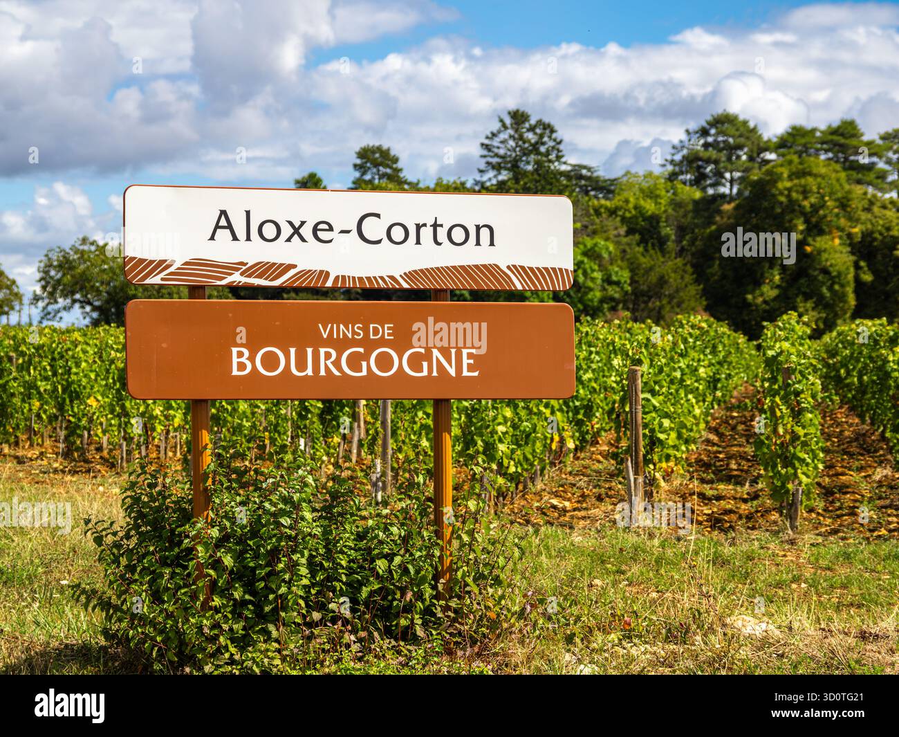 Ein Schild, das auf das Dorf Aloxe-corton in Burgund, Frankreich, hinweist, mit Weinbergen im Hintergrund unter teilweise bewölktem Himmel Stockfoto