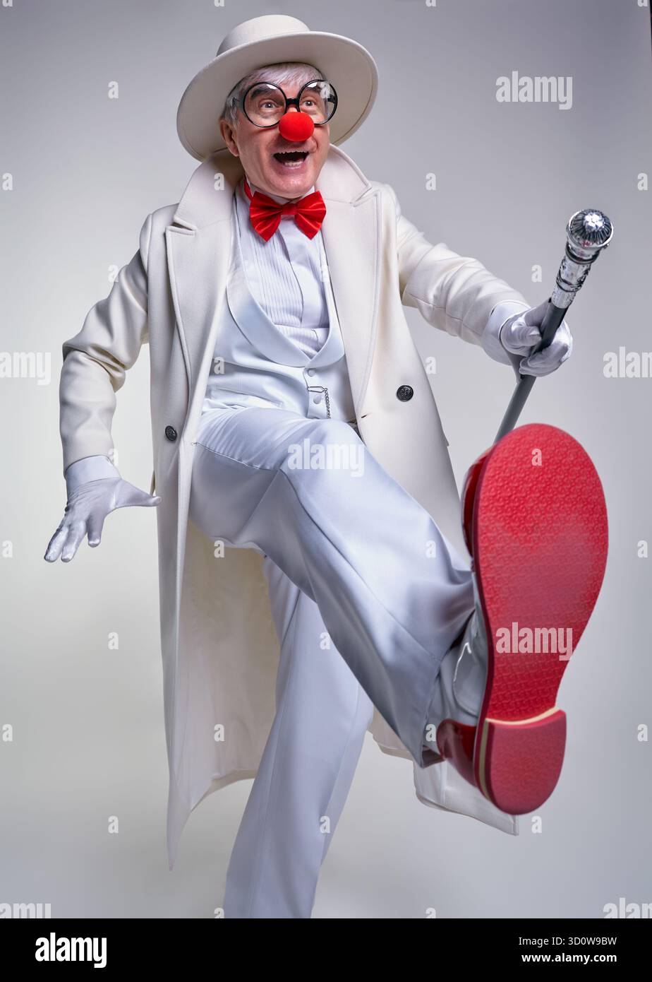 Ein älterer Clown in einem weißen Mantel und Hut mit Gehstock. Сlown tritt mit einem fröhlichen Gesichtsausdruck nach vorne. Der Mann trägt einen großen roten Papa Stockfoto