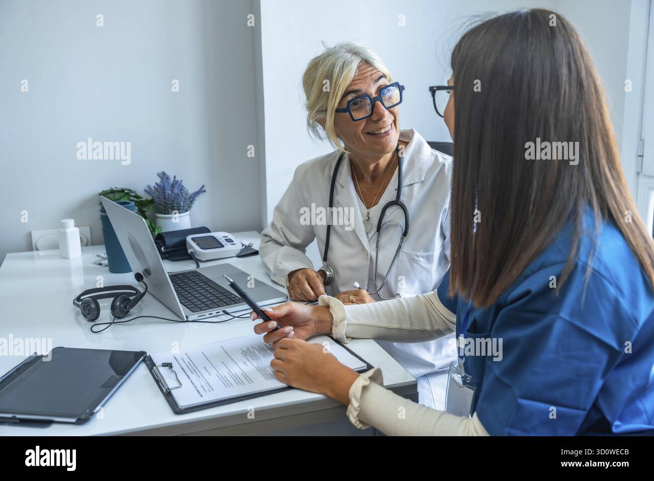 Zwei medizinische Fachkräfte diskutieren wichtige medizinische Dokumente und verwenden einen Laptop in einer Klinik, die Teamarbeit und umfassende Medizin repräsentieren Stockfoto
