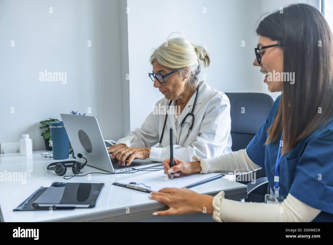 Zwei medizinische Fachkräfte, ein leitender Arzt, der auf einem Laptop tippt, und eine jüngere Krankenschwester, die Notizen auf einem Klemmbrett macht, arbeiten in einem modernen Gesundheitswagen zusammen Stockfoto