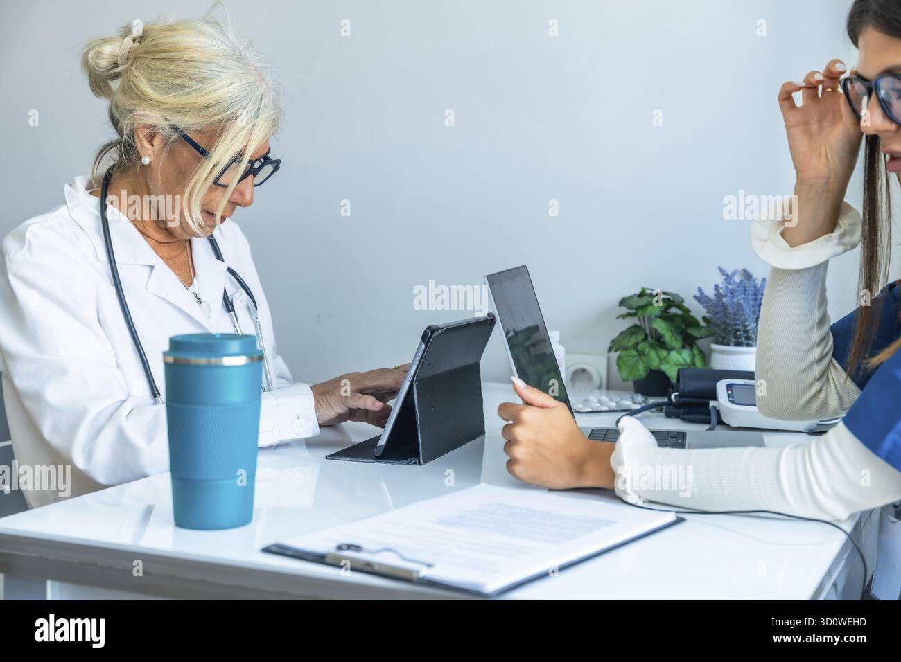 Zwei medizinische Fachkräfte arbeiten an einem Schreibtisch zusammen und nutzen digitale Tablets und Laptops für die Gesundheitsverwaltung Stockfoto