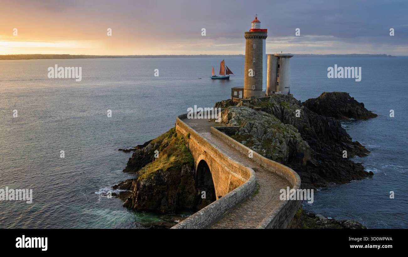 Ein malerischer Blick fängt den Leuchtturm Phare du Petit Minou ein, der stark auf einer felsigen Klippe steht. Ein Segelboot gleitet in der ruhigen See unter einem warmen Sonnenuntergang Himmel Stockfoto