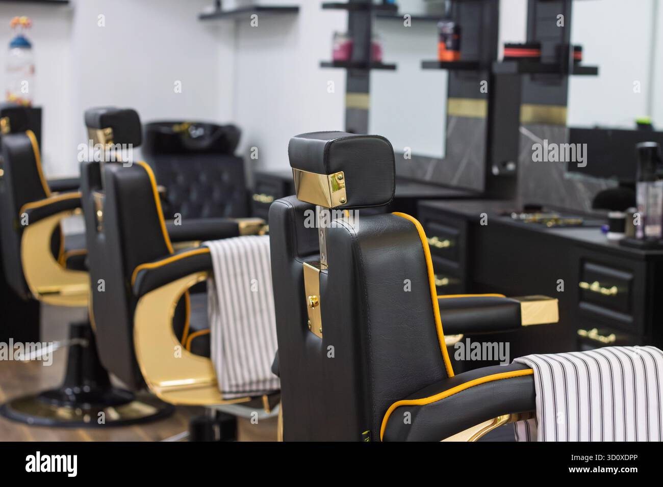 Rückansicht der schwarz-goldenen Barbershop-Stühle mit einem gestreiften Barber cape über dem Arm, das eine saubere und stilvolle Innenausstattung des Barbershops zeigt. Stockfoto