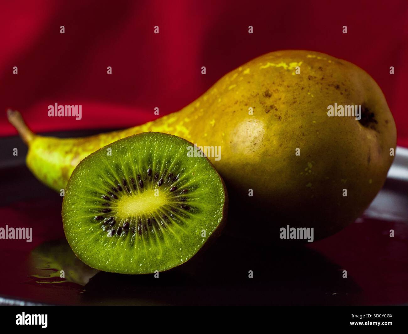 Frisches grünes Kiwi, das neben einer ganzen Reifen Birne auf einem dunklen Teller halbiert wird Stockfoto