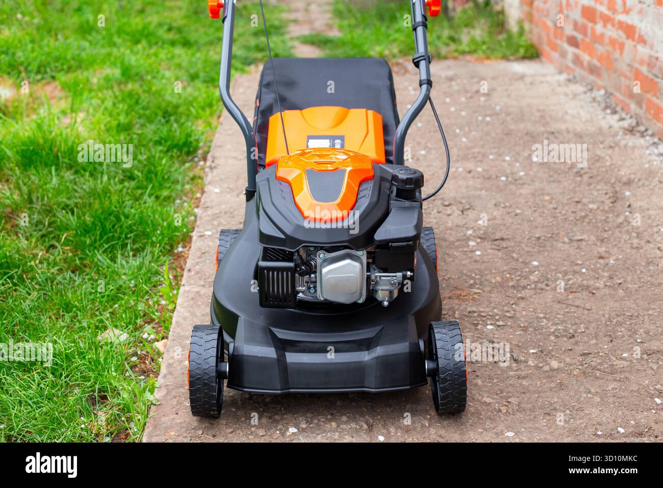 Ein moderner Rasenmäher in Schwarz und Orange befindet sich auf einer Betonterrasse neben einem grünen Gras und ist einsatzbereit. Stockfoto