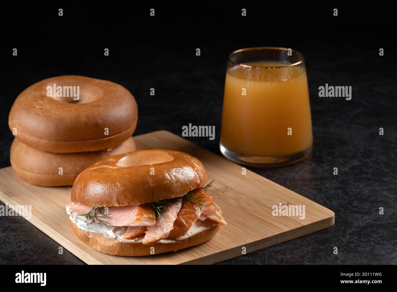 Ein Bagel mit atlantischem Lachs und ein Glas Saft liegen auf einem Holzbrett. Das Sandwich ist ein geräucherter Lachs-Bagel mit Frischkäse und Dill. Zwei einfarbige B Stockfoto
