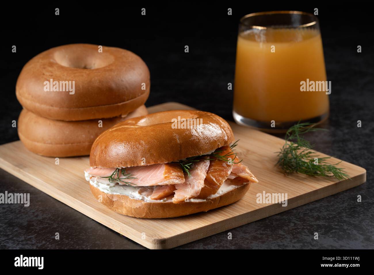 Ein Sandwich mit Lachs und Dill liegt auf einem Holzbrett neben einem Glas Saft. Das Sandwich wird in zwei Hälften geschnitten und auf eine andere Hälfte gelegt Stockfoto