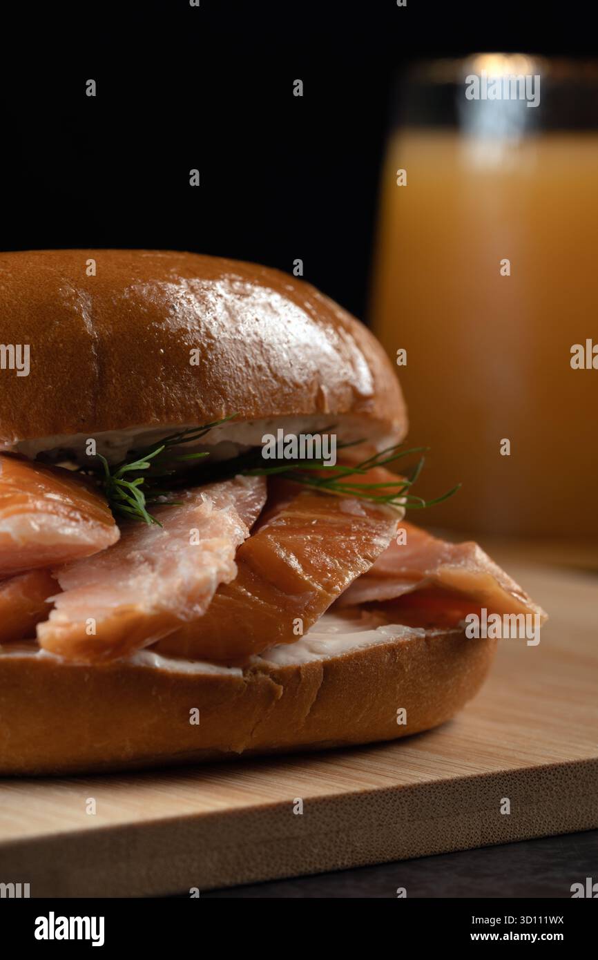 Ein Sandwich mit Lachs und Dill auf einem Brötchen sitzt auf einem hölzernen Schneidebrett. Ein Glas Orangensaft steht neben dem Sandwich Stockfoto