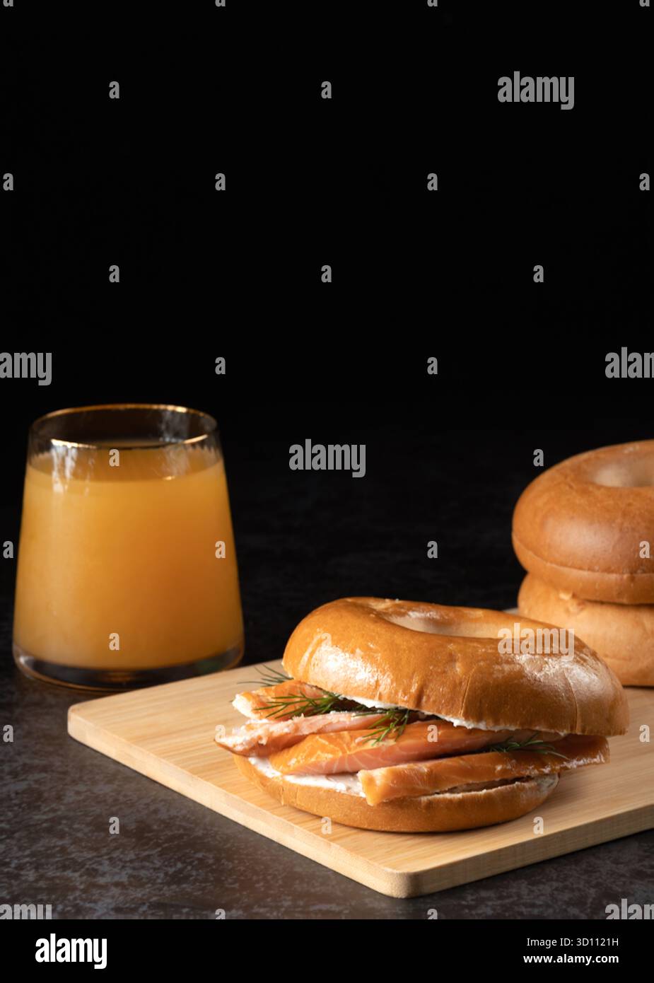 Ein Bagel und ein Glas Saft stehen auf einem Holztisch. Das Sandwich ist ein Räucherlachs-Bagel mit Dill-Gurken. Die Szene ist lässig und entspannt Stockfoto