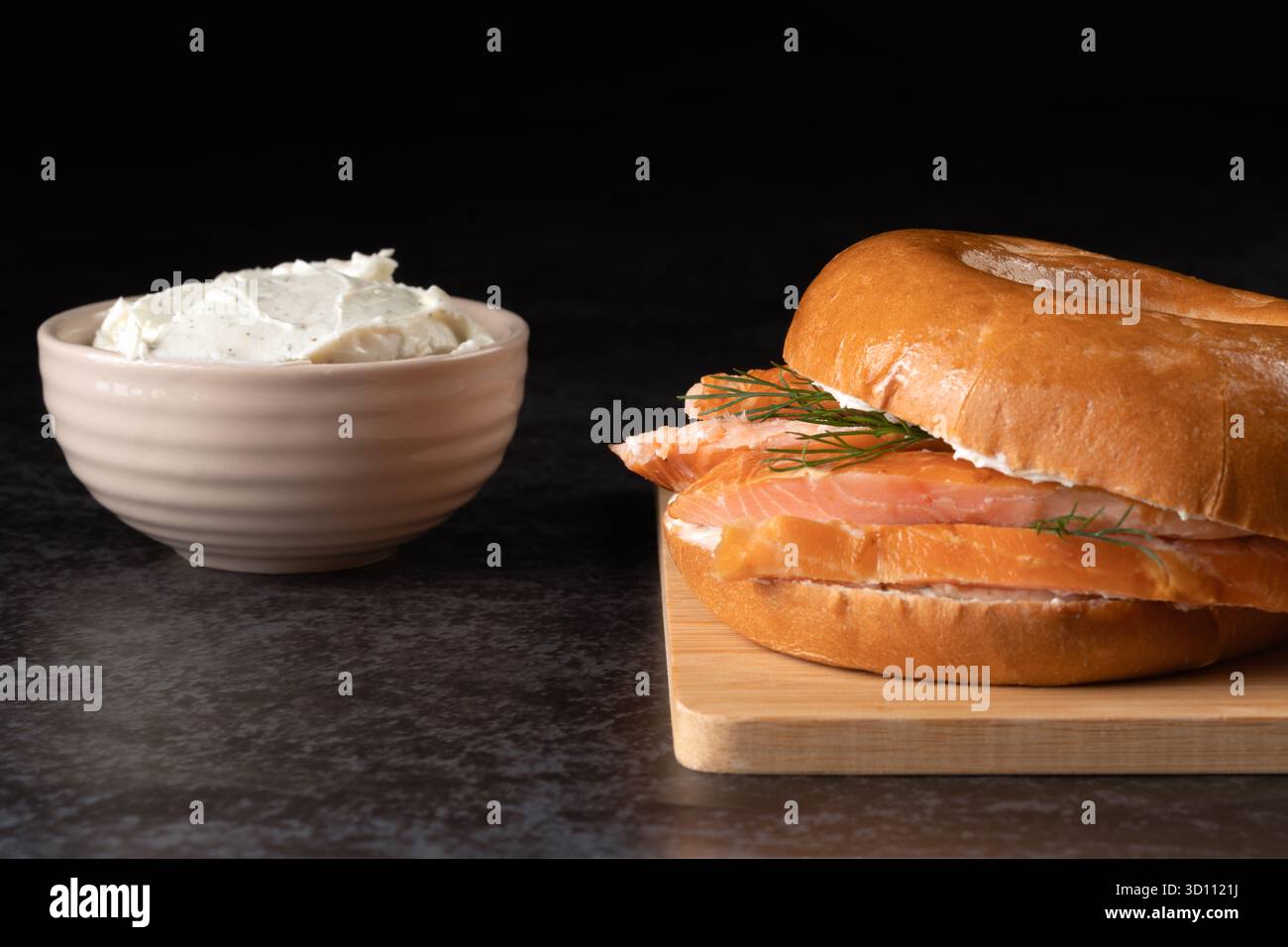 Ein Sandwich mit Lachs und Dill liegt auf einem Holzbrett. Eine Schüssel Frischkäse ist neben dem Sandwich Stockfoto
