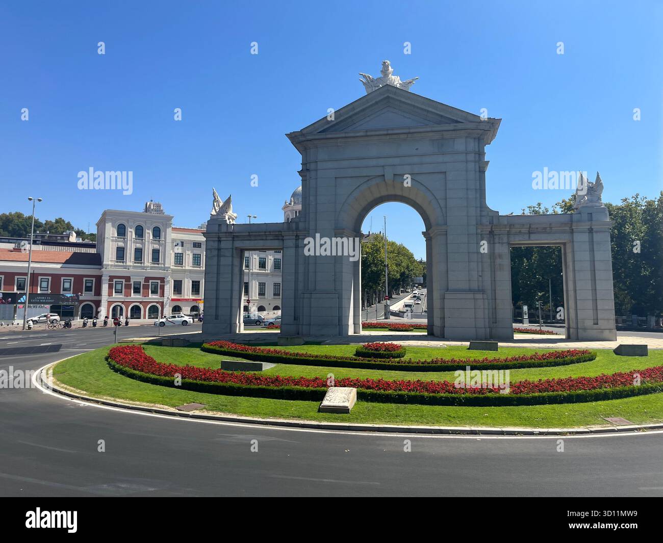 Seitenansicht der Puerta de Toledo in Madrid, Spanien, umgeben von roten Sommerblumen mit der Station Príncipe Pío links an einem sonnigen Tag. - Smartphone-aufgenommenes Stockfoto