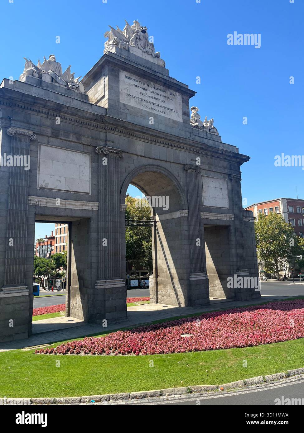 Seitenansicht der Puerta de Alcalá in Madrid, Spanien, umgeben von rosa Sommerblumen an einem sonnigen Tag. - Smartphone-aufgenommenes Stockfoto