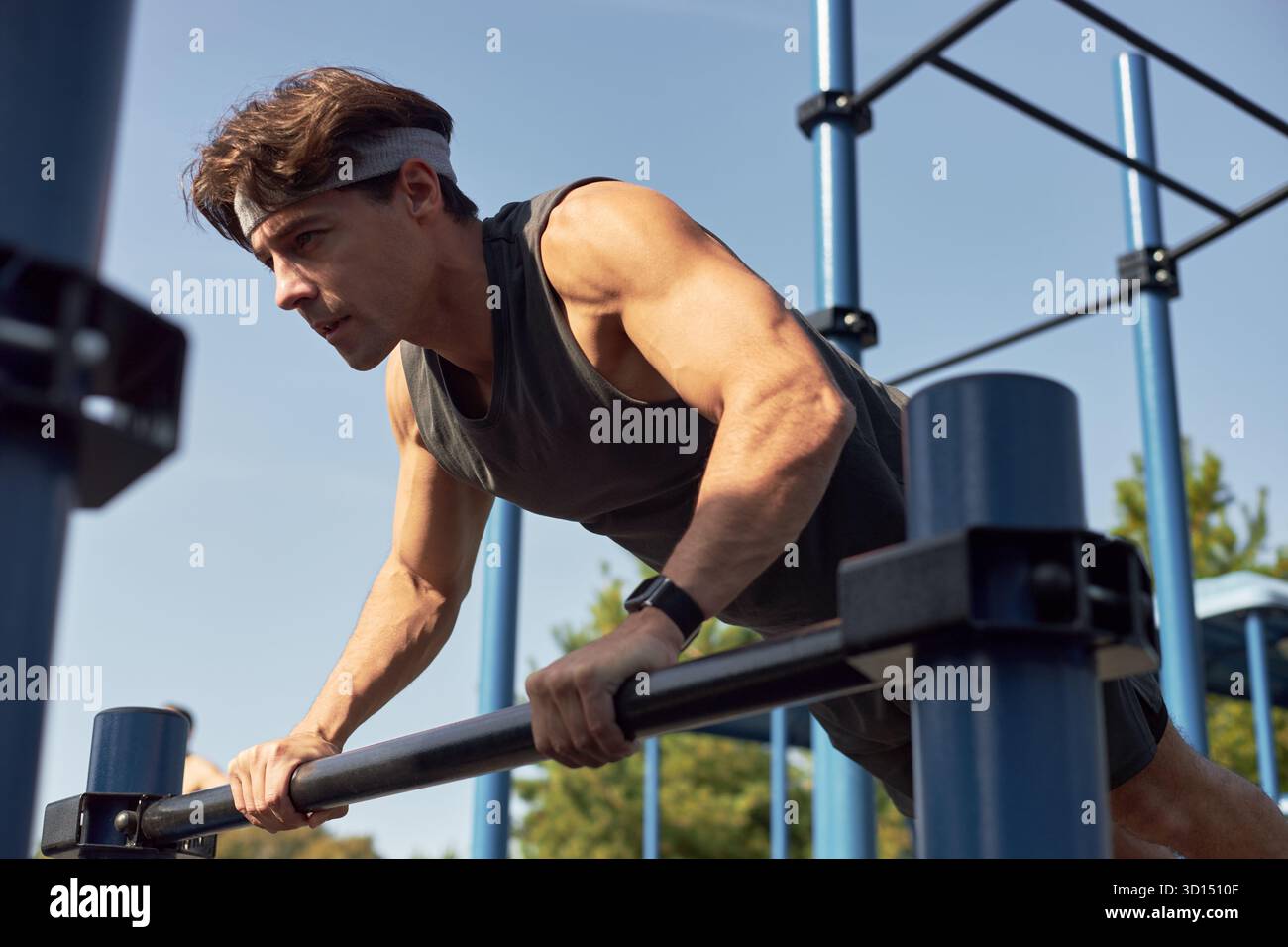 Kaukasischer junger erwachsener Mann, der im Freien Liegestütze auf parallelen Bars ausführt, Fitness-Tracker trägt, der sich auf das Training konzentriert, Muskeln engagierte sportliches Training im Outdoor-Fitnessstudio Stockfoto