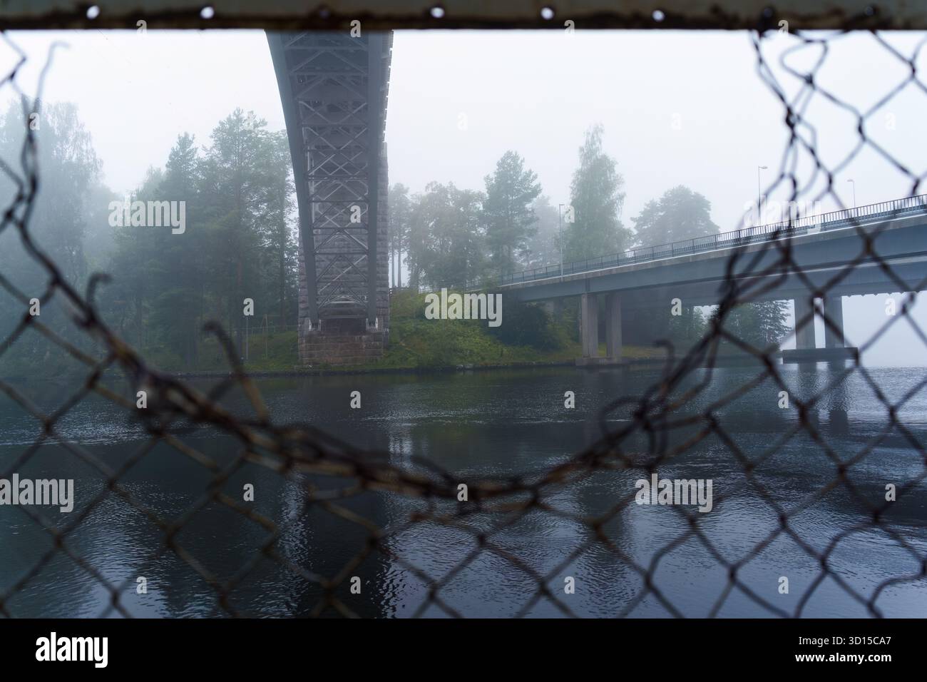Historische Heinola-Eisenbahnbrücke durch eine Lücke in einem Maschendrahtzaun an einem nebeligen Morgen. Stockfoto