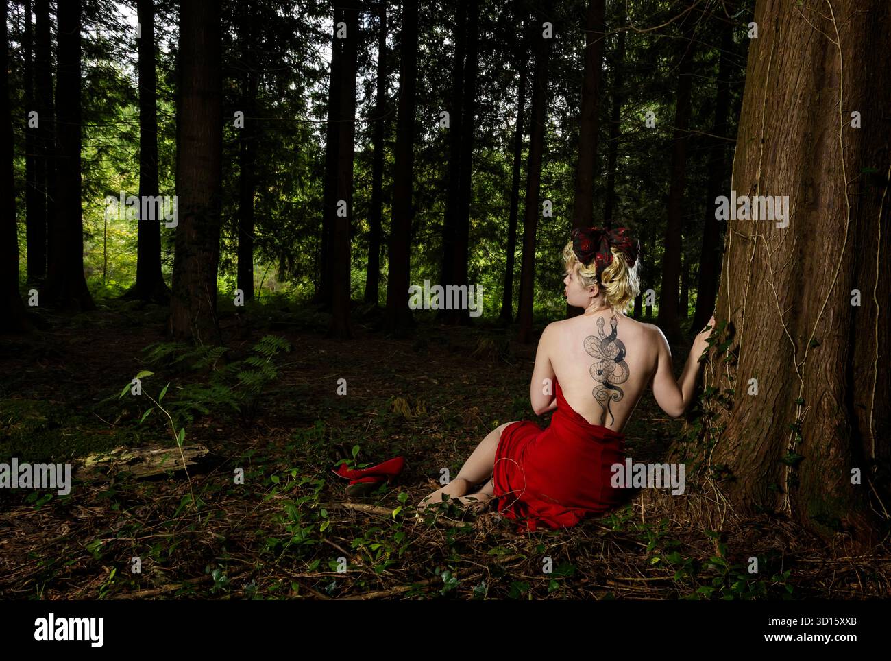 Eine schöne blonde Frau in einem roten Kleid, die im Wald sitzt und ihr Schlangentattoo auf dem Rücken zeigt. Stockfoto