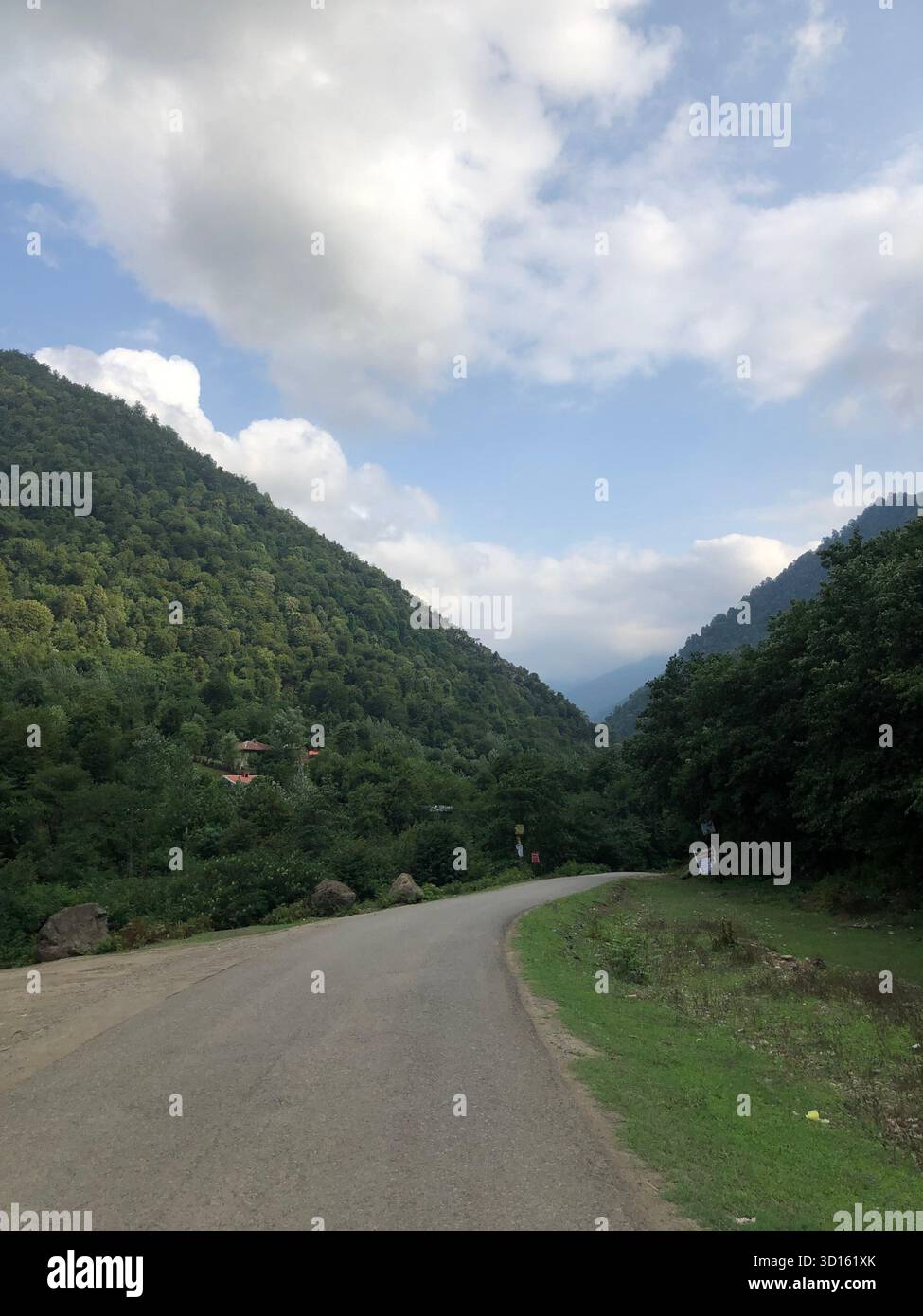 Eine gewundene Straße durch die üppigen hyrkanischen Wälder von Gilan, Iran; grüne Hänge steigen zu einem wolkenbewachsenen blauen Himmel. Stockfoto