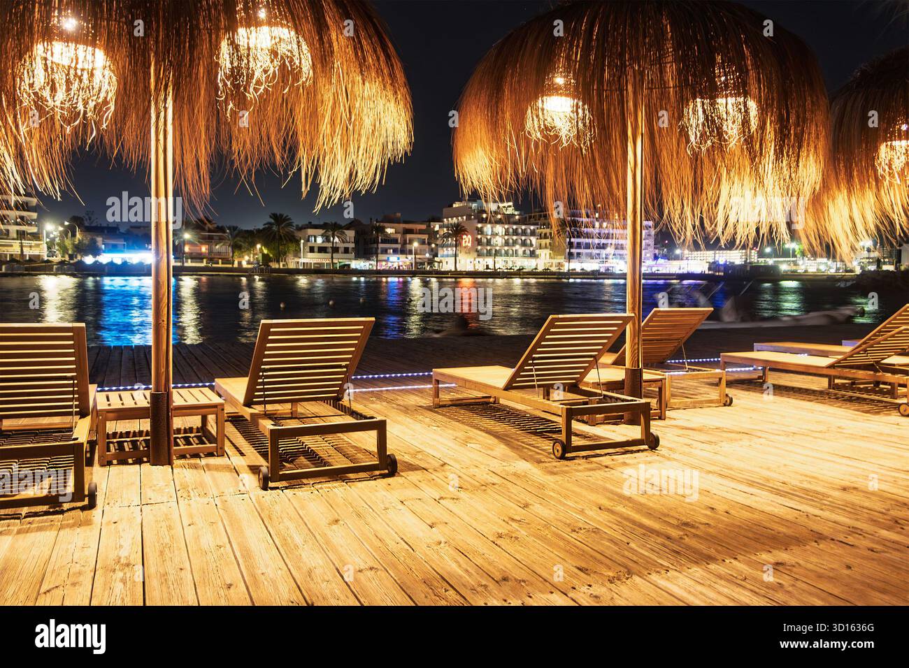 Leere hölzerne Sonnenliegen unter Strohschirmen auf einer Terrasse am Wasser bei Nacht mit Lichtern, die auf dem Wasser c'an pastilla mallorca reflektieren Stockfoto