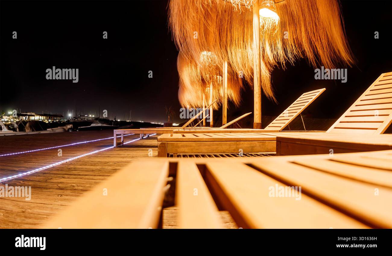 Leere hölzerne Liegestühle auf einer Promenade, die nachts von warmen tropischen Lichtern beleuchtet wird, in der Nähe einer Marina Bucht mit Booten und ruhigen Gewässern c'an pastilla mallorca Stockfoto