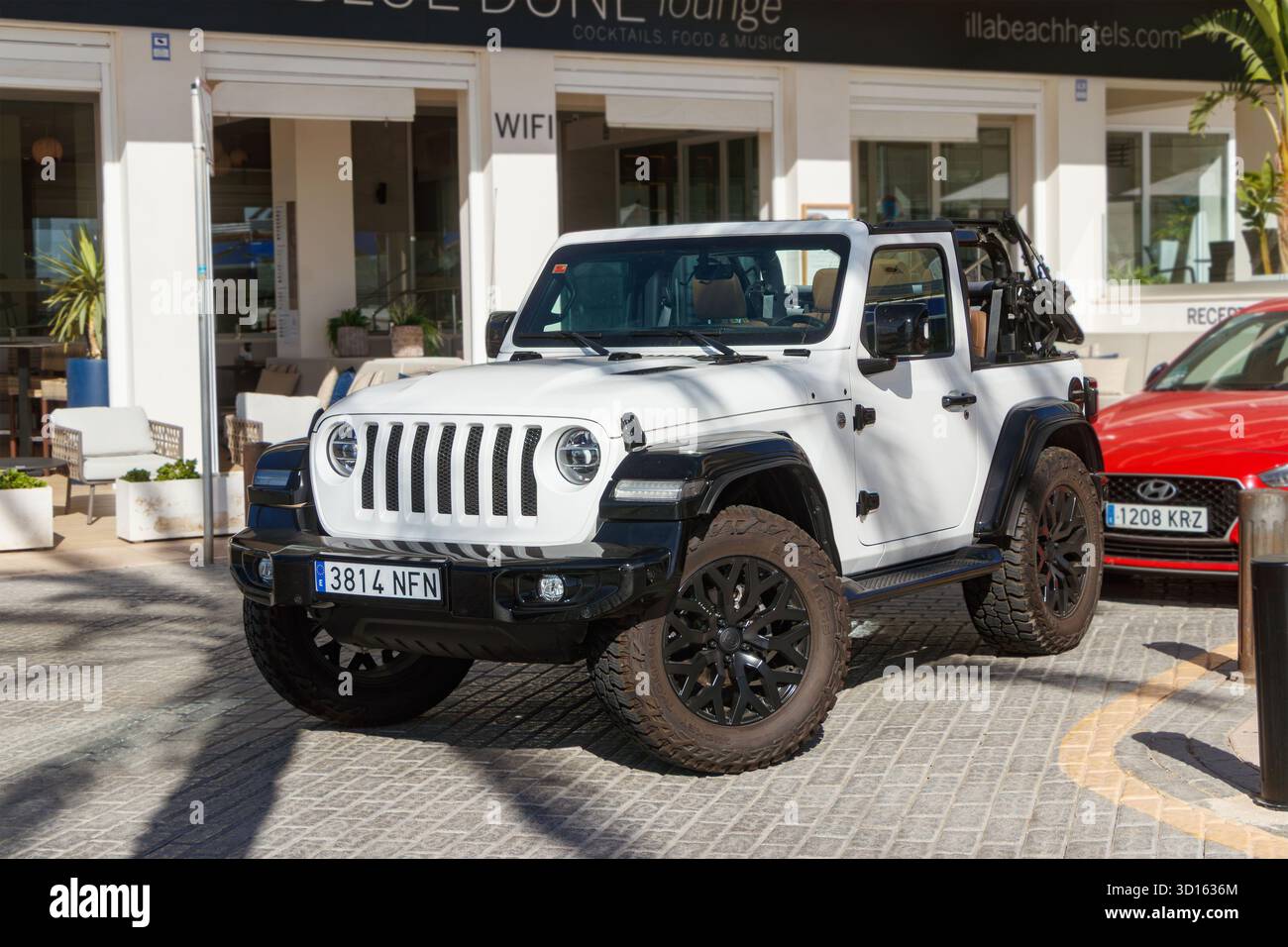 Weißer Jeep Wrangler parkte auf einer Kopfsteinpflasterstraße in der Nähe eines modernen Cafés und Hoteleingangs in einem sonnigen Küstengebiet Stockfoto