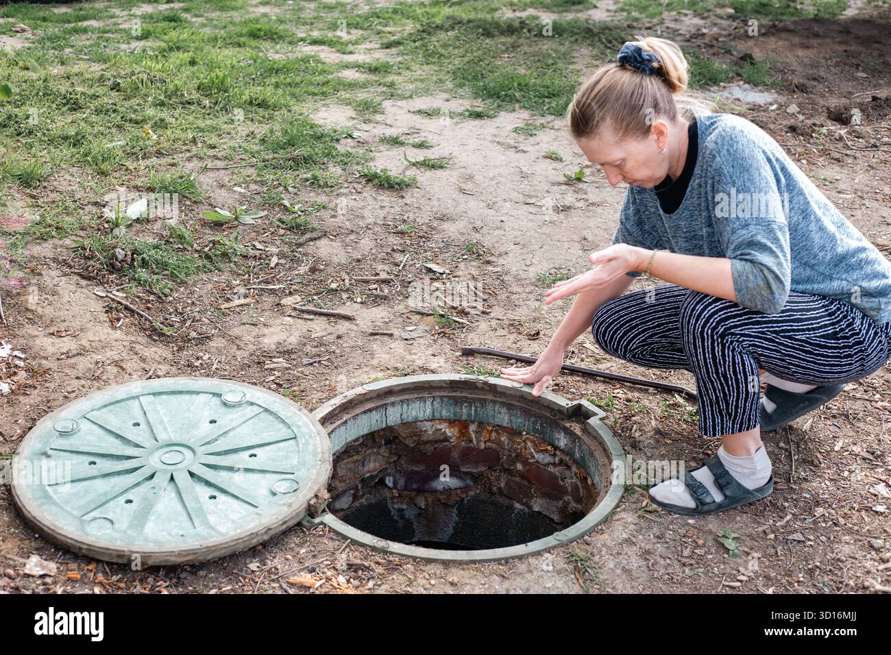 Eine Frau lehnt sich über ein offenes Mannloch, überprüft eine Klärgrube und den Stand des Abwassers, das aus dem unterirdischen Kanalisationssystem abgeleitet wird. Stockfoto