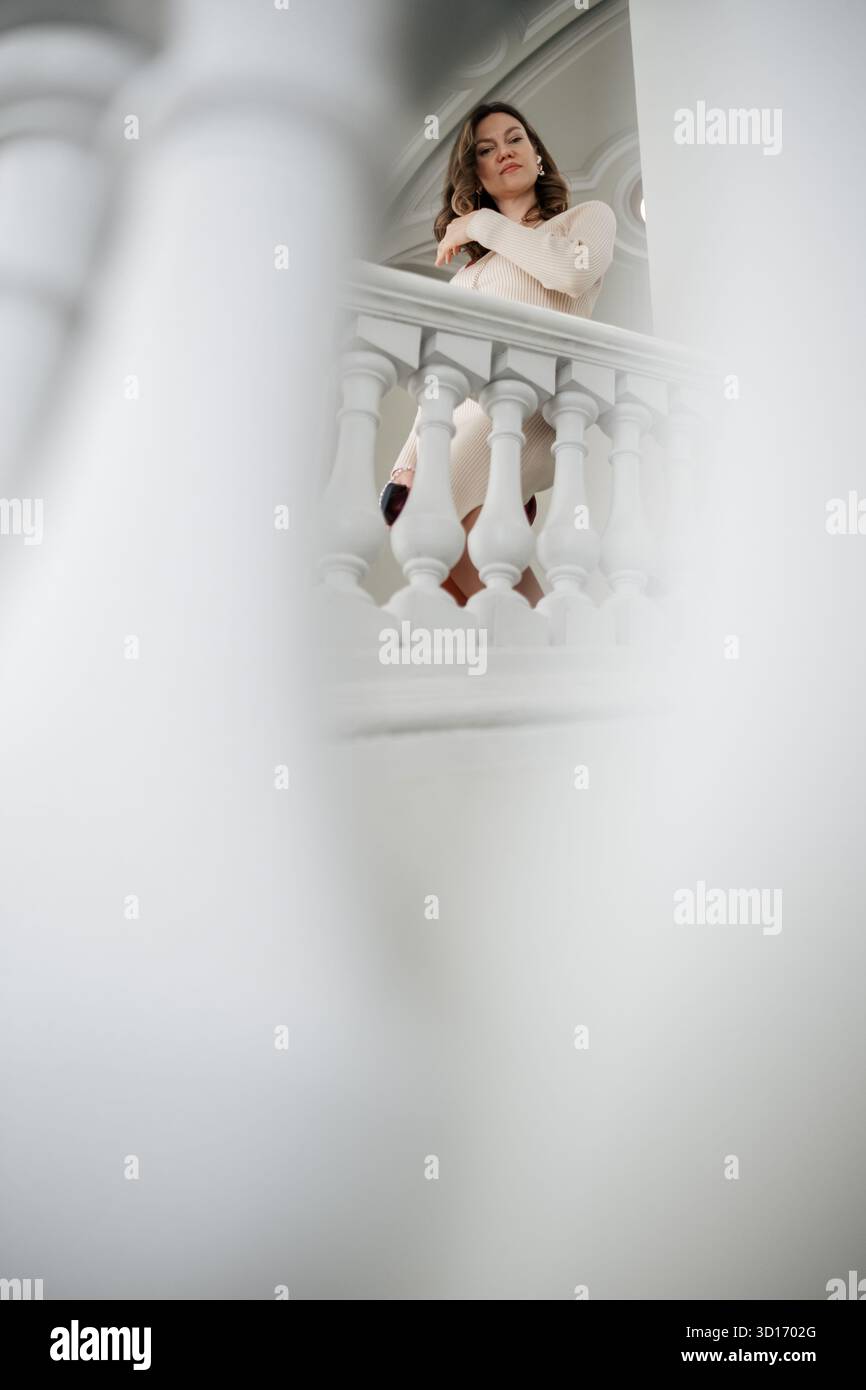 Eine Frau steht mit kontemplativem Ausdruck, ihre Arme auf einem weißen, verzierten Balkongeländer in einem sanft beleuchteten, eleganten Interieur. Stockfoto