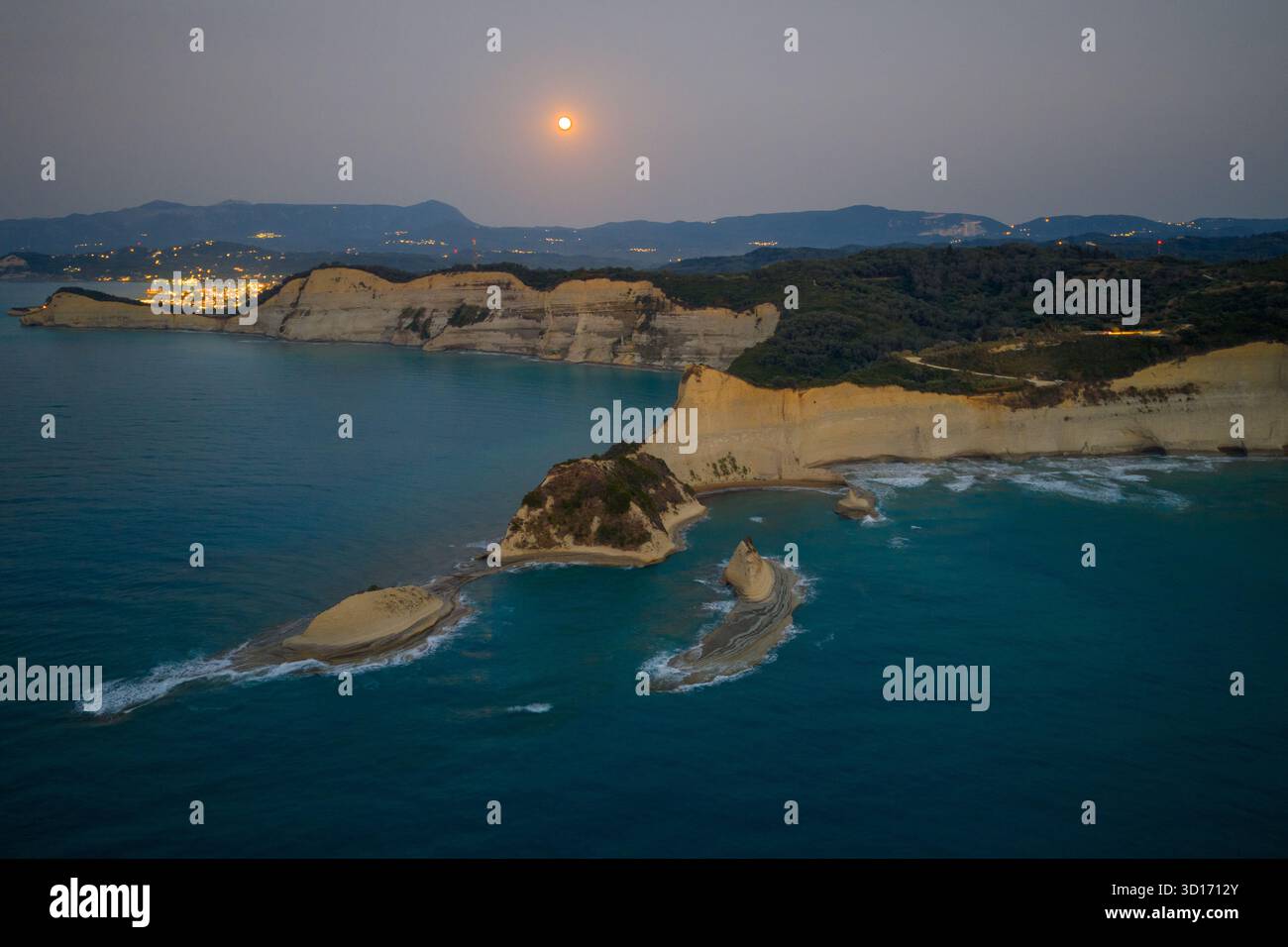 Majestätische Aussicht auf den Vollmond, der über den einzigartigen weißen Klippenformationen des Kap Drastis auf Korfu, Griechenland, aufsteigt, mit Lichtern der Stadt Stockfoto