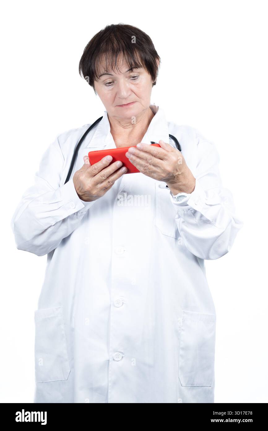 Leitende Ärztin mit weißem Labormantel und Stethoskop, konzentriert auf die Verwendung eines roten Smartphones, die Telemedizin, digitale Gesundheitsversorgung repräsentieren Stockfoto