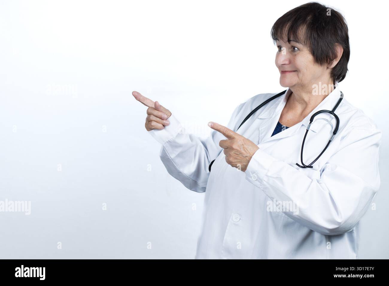 Lächelnde Seniorenärztin trägt einen weißen Labormantel und Stethoskop, zeigt mit beiden Händen, zeigt oder empfiehlt medizinische Informationen Stockfoto
