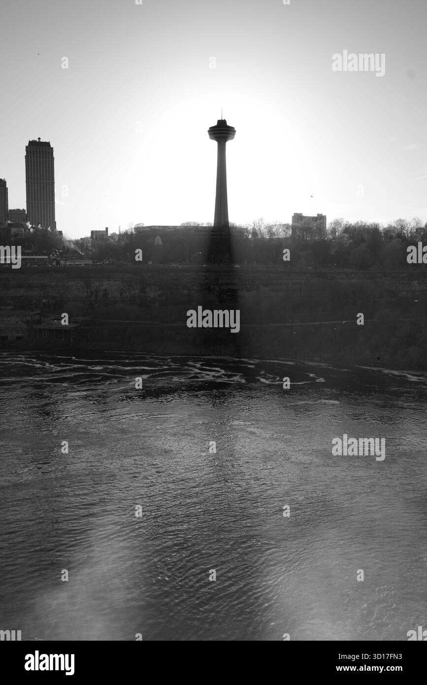Schwarzweiß-Foto des Skylon Tower von der amerikanischen Seite aus gesehen, von Sonnenlicht umgeben, während er über dem Niagara River und dem umliegenden Himmel aufsteigt Stockfoto
