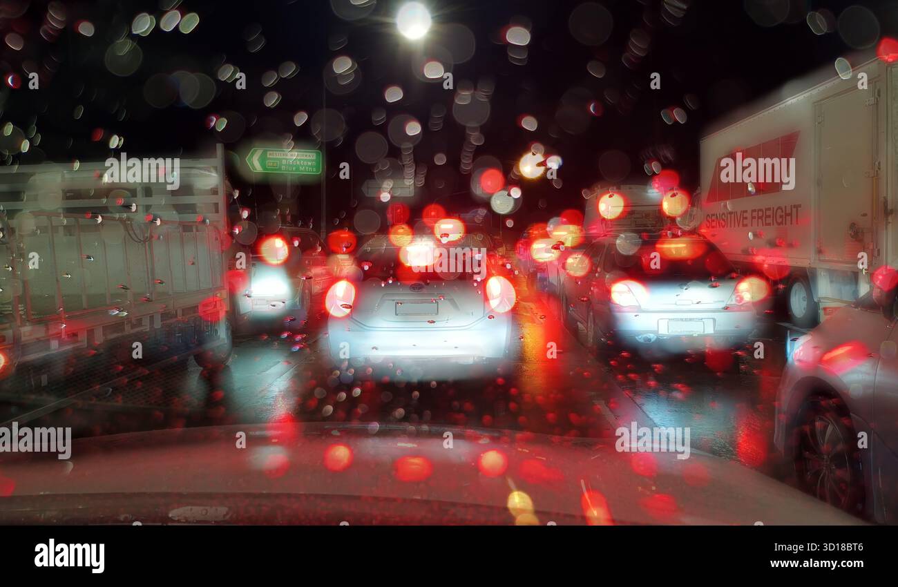 Verkehrsstau an der Kreuzung mit roten Rücklichtern von Lastwagen und Autos, gesehen durch verschwommene Regenstellen auf der Windschutzscheibe des Autos am frühen Morgen Sydney, Australien Stockfoto