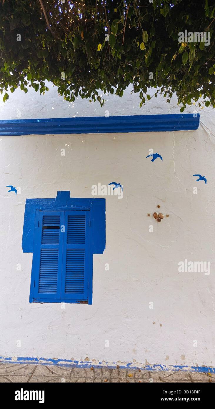 Traditionelle weiß getünchte Gebäudefassade mit leuchtend blau lackierten Rollläden, Zierleisten und dekorativen fliegenden Vögeln. Typisch mediterrane Architektur - Smartphone-aufgenommenes Stockfoto