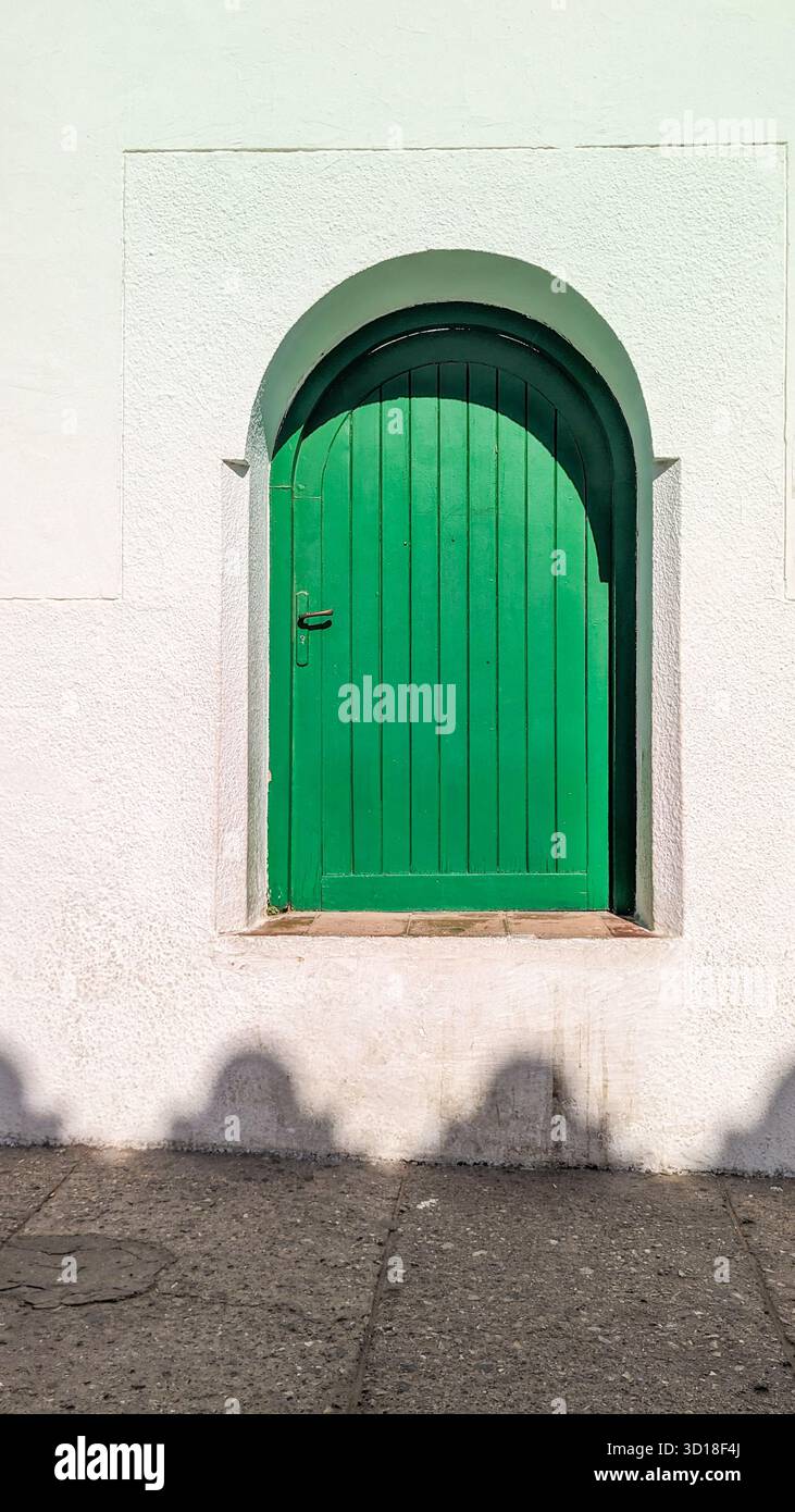 Leuchtend gewölbtes grünes Holzfenster in einer strukturierten weißen Succo-Wand mit starken Schatten auf dem Bürgersteig. - Smartphone-aufgenommenes Stockfoto