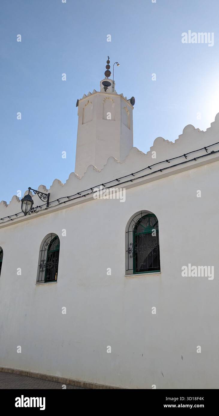 Minarett mit weißer Moschee und zinnenförmiger Attika mit Bogenfenstern und dekorativen Eisengittern vor klarem blauem Himmel Stockfoto