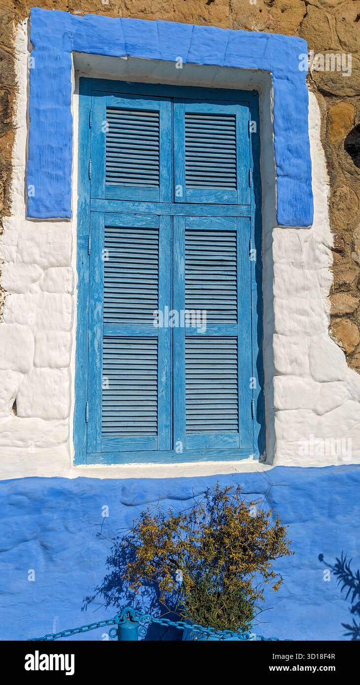 Fenster im traditionellen mediterranen Stil mit blauen Holzläden, weiß getünchter Steinwand, markanten blauen Zierleisten und grüner Pflanze, die einen natürlichen Touch verleiht - Smartphone-aufgenommenes Stockfoto